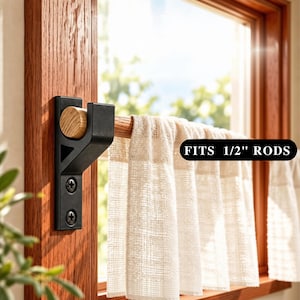 May include: A black metal curtain rod bracket mounted to a wooden window frame, holding a wooden rod with a light-colored fabric curtain. The bracket features a U-shaped cradle and two screws. A black label with white text reads FITS 1/2" RODS.