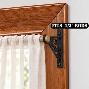 May include: A black metal curtain rod bracket with an ornate scrollwork design mounted on a wooden window frame. The bracket holds a light-colored wooden rod with a sheer white curtain. Text on the image states: FITS 1/2" RODS.