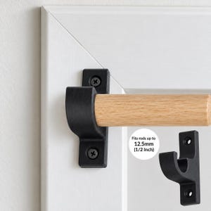 May include: A matte black metal curtain rod bracket mounted with two screws, holding a light-colored wooden rod. A separate bracket is shown to the right. Text on the image states: Fits rods up to 12.5mm (1/2 Inch).