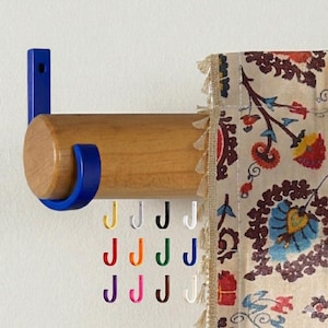 Puede incluir: Una barra de cortina de madera con soportes metálicos azules. Una cortina con estampado floral cuelga a la derecha. Debajo, hay ganchos en forma de J de colores en amarillo, blanco, negro, rojo, naranja, verde, morado, rosa, marrón y blanco.