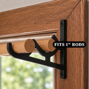 May include: A black metal double curtain rod bracket mounted to a wooden window frame, holding two light brown wooden rods. The bracket features a black label with white text that reads FITS 1" RODS, indicating the size capacity for the rods.