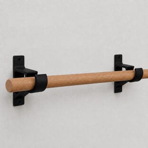 May include: A wall-mounted rod featuring a natural light brown wooden dowel supported by two matte black metal brackets. Each bracket is secured with visible screws, holding the horizontal wooden bar firmly in place for storage or display.