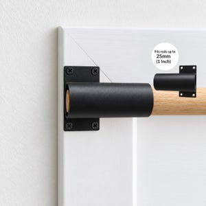 May include: A matte black metal curtain rod bracket with a rectangular mounting plate and four screw holes. The bracket holds a light-coloured wooden rod. Text on the image states: Fits rods up to 25mm (1 Inch).