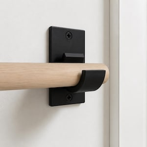 May include: A light-colored wooden rod held by a matte black metal wall bracket. The bracket features a rectangular base plate secured with two visible screws and a curved hook that supports the horizontal rod in a fixed position.
