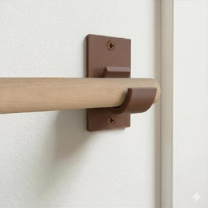 Può includere: Un bastone in legno di colore chiaro è sostenuto da una staffa da parete rettangolare di colore marrone. La staffa presenta un supporto curvo per sostenere il bastone ed è fissata alla parete con due viti a croce visibili, una in alto e una in basso.