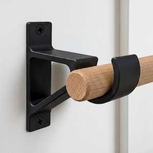 May include: A wall-mounted bracket featuring a matte black finish with two visible screws. The bracket supports a cylindrical light-toned wooden rod that rests within a curved, black metal cradle extending from the main support structure.