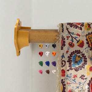 May include: A gold-colored curtain rod bracket and wooden rod are mounted on a white wall. A patterned curtain with floral designs hangs from the rod. A selection of colorful, heart-shaped curtain tiebacks are displayed.
