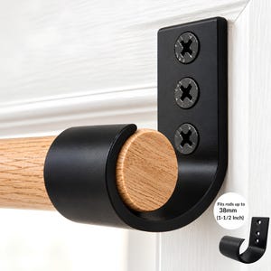 May include: A matte black metal curtain rod bracket mounted with three screws, holding a light-colored wooden rod. The bracket features a curved cradle design and includes text stating it fits rods up to 38mm (1-1/2 inch).