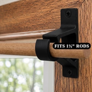 Puede incluir: Un soporte de metal negro mate para barra de cortina montado en un marco de ventana de madera, sosteniendo una barra de madera clara. El soporte tiene una base en forma de U y dos orificios para tornillos. Una etiqueta negra dice FITS 1 1/2" RODS.