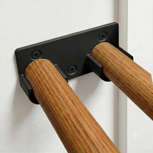 May include: A wall-mounted hardware fixture featuring a rectangular matte black metal base plate secured with two visible screws. Two cylindrical wooden dowels with a natural light brown wood grain finish extend outward from the black metal brackets.