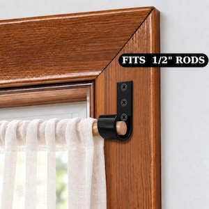 May include: A black metal curtain rod bracket mounted to a wooden window frame with three screws, holding a light-colored wooden rod with a sheer white curtain. The bracket features a label that reads FITS 1/2" RODS for clear sizing reference.