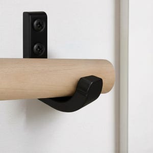 Puede incluir: Un conjunto de herrajes de pared con una barra de madera de tono claro sujeta por un soporte de metal negro mate. El soporte está fijado con dos tornillos negros visibles e incluye un gancho negro curvado bajo la barra horizontal.