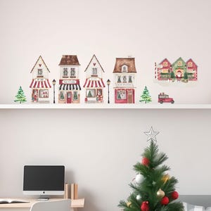 May include: A watercolour illustration of a Christmas village scene. The image shows several buildings with red and white striped awnings, Christmas trees, and a red truck. The buildings are arranged on a white shelf, with a decorated Christmas tree in the foreground.