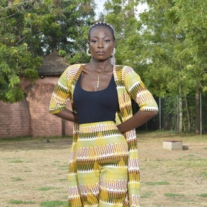 May include: A woman wearing a yellow, green, and red patterned pantsuit with a black tank top underneath. She is wearing white sandals and a gold necklace.