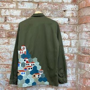 May include: A green long-sleeved shirt with a patchwork design of a village on the back. The design features houses with red roofs and white walls, and green trees.