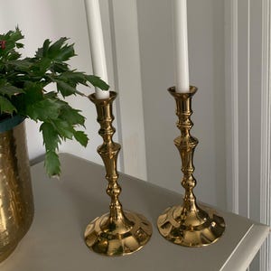 May include: Two gold-colored candle holders with white candles. The candle holders have a detailed design and a round base. A green plant in a gold pot is visible on the left side of the image.