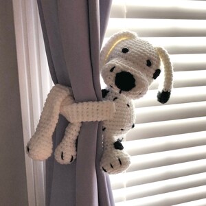 May include: A white crocheted dog with black spots is holding a curtain open. The dog is a cute and playful design, perfect for a child's room.