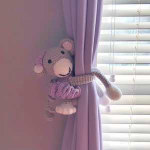 May include: A crocheted monkey toy in gray and purple is holding a lavender curtain tieback. The monkey is holding the tieback with its arms and legs, and its head is peeking out from behind the curtain.