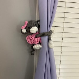 May include: A gray crocheted monkey doll wearing a pink tutu is holding onto a lavender curtain. The monkey has a white face and black eyes.