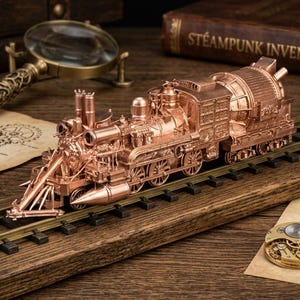 Puede incluir: Una locomotora de vapor modelo de metal color cobre se encuentra sobre una vía de tren en miniatura. El tren presenta detalles mecánicos intrincados, incluyendo ruedas, pistones y una gran caldera trasera, junto a una lupa y un libro titulado Steampunk Inventions.