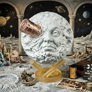 May include: A white, textured moon sculpture with a human face and a copper-colored rocket ship embedded in its eye. The sculpture rests on a translucent yellow stand, surrounded by a film canister, a clapperboard, and a vintage compass.