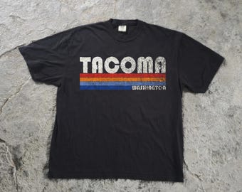 May include: A black short-sleeved t-shirt featuring the word TACOMA in large white distressed lettering above horizontal stripes in red, orange, and blue, with the word WASHINGTON printed in white below the stripes.