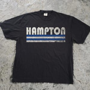 May include: A black short-sleeved t-shirt featuring a distressed white graphic that reads HAMPTON in large capital letters. Below the text are two horizontal stripes, one blue and one white, with the word VIRGINIA printed in small white letters.