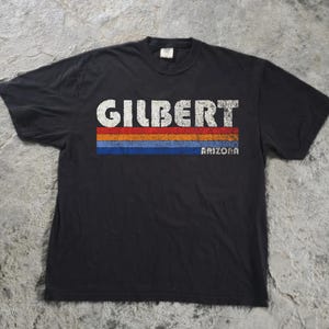 May include: A black short-sleeve t-shirt featuring the white text GILBERT above three horizontal stripes in red, orange, and blue. The word ARIZONA is printed in white to the right of the blue stripe. The graphic has a distressed texture.
