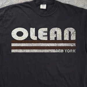 May include: A black short-sleeve t-shirt featuring the word OLEAN in large, distressed white capital letters. Below the text are two horizontal brown stripes and two horizontal white stripes, with the words NEW YORK in white.