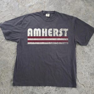 May include: A charcoal grey short-sleeve t-shirt featuring the word AMHERST in distressed white capital letters above two horizontal maroon stripes and the word MASSACHUSETTS in smaller white capital letters on the bottom right side.