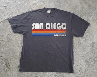May include: A charcoal gray short-sleeve t-shirt featuring the white distressed text SAN DIEGO above three horizontal stripes in red, orange, and blue. The word CALIFORNIA is printed in small white letters below the blue stripe.