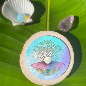 May include: A round wooden coaster with a blue and purple gradient background featuring a white pearl inside a pink and white seashell. The text "Shell Yeah" is written in white cursive letters on the coaster. The text "Pearl & Stone Illustrations" is written in white at the bottom of the coaster.