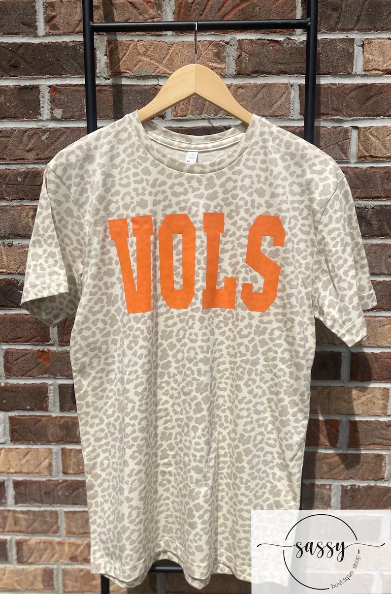 May include: A beige t-shirt with a leopard print pattern and the word "VOLS" in orange block letters. The shirt is hanging on a wooden hanger against a brick wall background. The shirt is from the "Sassy Boutique Shop".