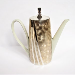 Könnte beinhalten: Eine silber- und cremefarbene Kaffeekanne mit Deckel und gebogenem Griff. Die Kanne hat eine strukturierte, gehämmert Metalloberfläche am Hauptkörper mit vertikalen weißen Streifen. Ausguss und Griff sind cremefarben.