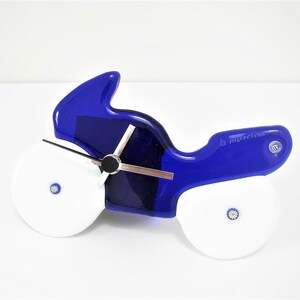 Könnte beinhalten: Eine blaue Glas-Motorrad-Uhr mit weißen Rädern und einem silbernen Zifferblatt. Die Uhr hat ein kleines Blumenmuster auf jedem Rad und die Worte "la murrina" an der Seite.