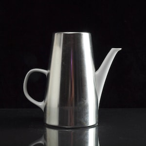 Könnte beinhalten: Eine moderne Kaffeekanne aus Edelstahl mit weißem Ausguss und Griff. Die Kanne hat eine konische Form und eine reflektierende Oberfläche, vor dunklem Hintergrund. Der Griff ist gebogen und der Ausguss ist schlank.