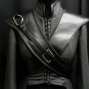 May include: A grey jacket with a high collar and black leather detailing. The leather includes a shoulder piece and a sash across the chest, decorated with metal studs and a large ring. The jacket is set against a dark backdrop.