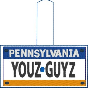 Könnte beinhalten: Ein blauer und weißer Anhänger in Form eines Pennsylvania-Kennzeichens mit dem Text "YOUZ-GUYZ" und einem kleinen blauen Staatssymbol.