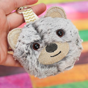 May include: A gray plush keychain with a brown embroidered face and black eyes. The keychain is attached to a gold and white striped ribbon.