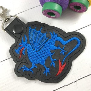 May include: A black keychain with a silver clasp and a dragon design. The dragon is blue with red accents and embroidered on a black background. The dragon has wings and a long tail.
