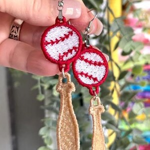 Op de afbeelding: Een paar oorbellen met rode en witte baseball-charms. Een charm is een baseball met witte stiksels en de andere is een bruine honkbalknuppel.