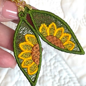 May include: A pair of green and yellow sunflower earrings with gold hooks. The earrings are made of fabric and have a delicate, lace-like design.
