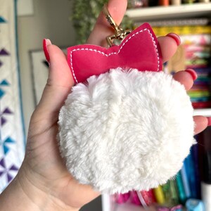 Puede incluir: Un llavero de pompón blanco esponjoso con un lazo rosa y un anillo de metal dorado.