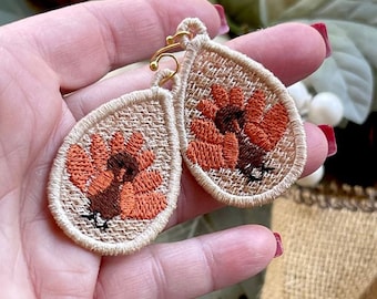 May include: A pair of beige fabric earrings with embroidered orange and brown turkeys. The earrings are teardrop shaped and have gold-colored hooks.