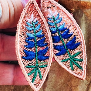 May include: Embroidered leaf-shaped earrings with a bluebell design. The earrings have a peach-coloured, openwork border and a blue, green, and white floral pattern. The earrings are suspended from a small, delicate hook.