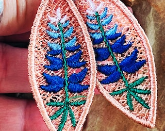 Könnte beinhalten: Gestickte ohrringförmige Blätter mit einem Lupinen-Design. Die Ohrringe haben einen pfirsichfarbenen, durchbrochenen Rand und ein blaues, grünes und weißes Blumenmuster. Die Ohrringe sind an einem kleinen, filigranen Haken aufgehängt.
