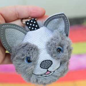 May include: A gray and white plush keychain with a black and white polka dot ribbon. The keychain is shaped like a dog's head with embroidered eyes, nose, and mouth.