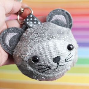 Puede incluir: Un llavero de ratón de peluche gris con ojos negros y bigotes. El ratón tiene un acabado brillante y está unido a una cinta de lunares negros y blancos.