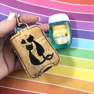 Könnte beinhalten: Kork-Handdesinfektionsmittel-Halter mit einem schwarzen Katzenmotiv. Der Halter ist an einem silbernen Schlüsselanhänger befestigt. Neben dem Halter befindet sich eine kleine Flasche Pocketbac-Handdesinfektionsmittel. Die Flasche ist blau und trägt die Aufschrift "At the Beach" auf dem Etikett.