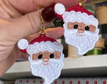 May include: A pair of gold earrings with embroidered Santa Claus faces. The Santa Claus faces are white with red hats and white beards.